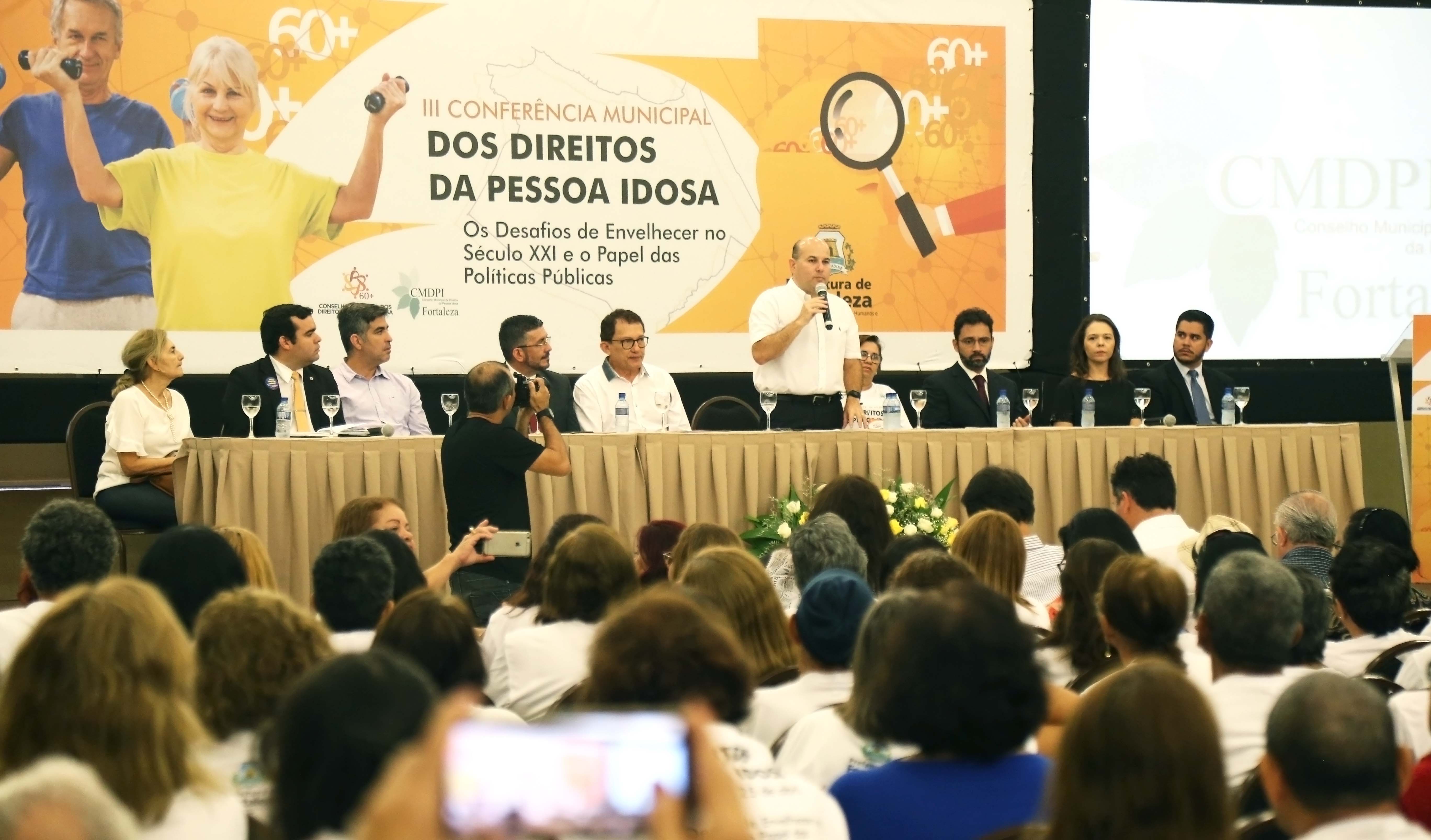 Mesa de abertura da Conferência Municipal dos Direitos da Pessoa Idosa com várias pessoas sentadas e o prefeito roberto cláudio em pé, discursando